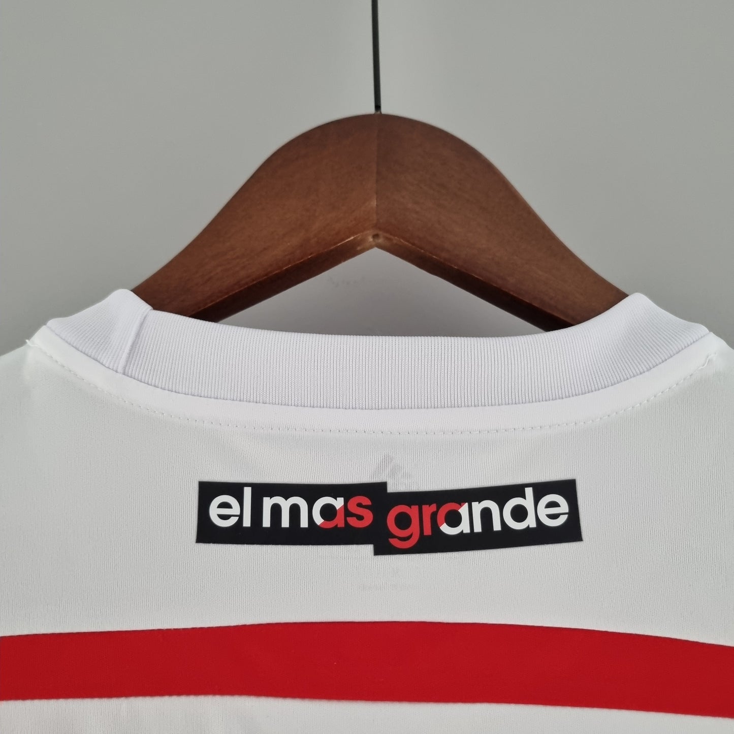 Camiseta de fútbol blanca con la frase 'el mas grande' en la parte trasera, sobre percha de madera
