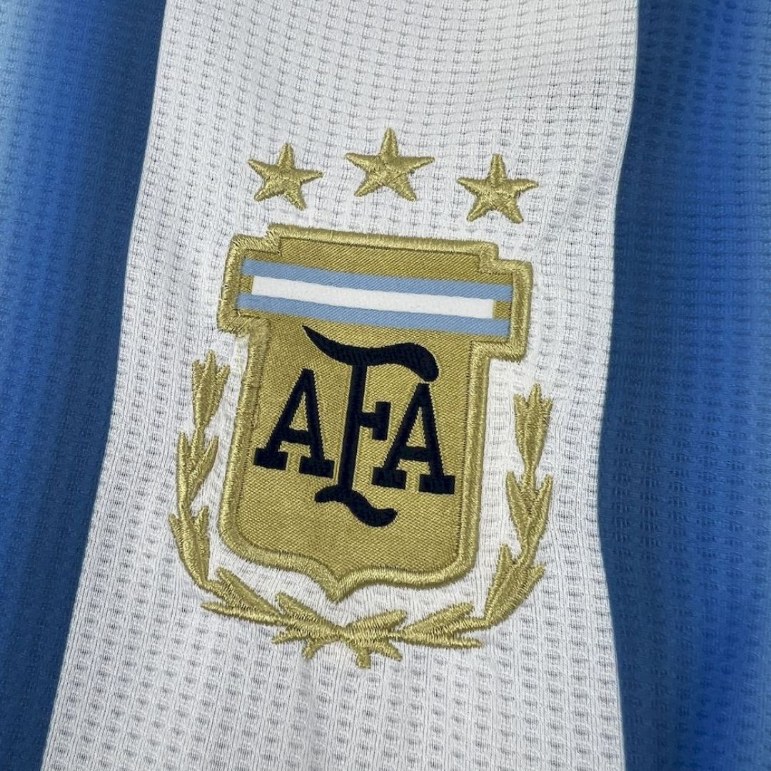 Escudo de la AFA con tres estrellas en camiseta Argentina 2026, fondo blanco y celeste