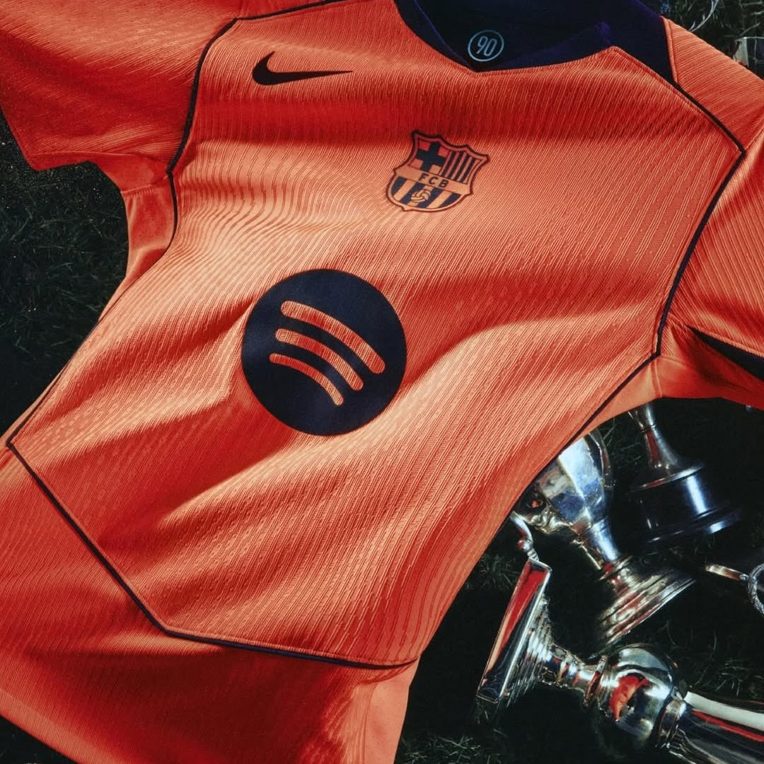 Camiseta nueva FC Barcelona naranja con logo Nike y Spotify, sobre césped y trofeos, Reino del Futbol
