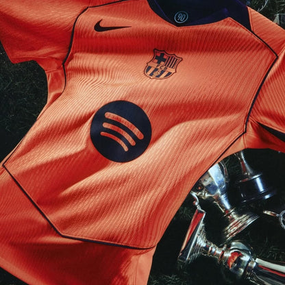 Camiseta nueva FC Barcelona naranja con logo Nike y Spotify, sobre césped y trofeos, Reino del Futbol