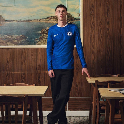 Hombre con camiseta azul Chelsea 2025, en cafetería, Reino del Futbol, fondo costero.