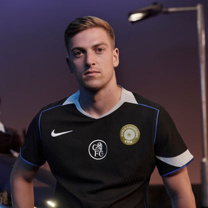 Aficionado luce camiseta negra Chelsea Third Kit con escudo FIFA, fondo urbano al atardecer.