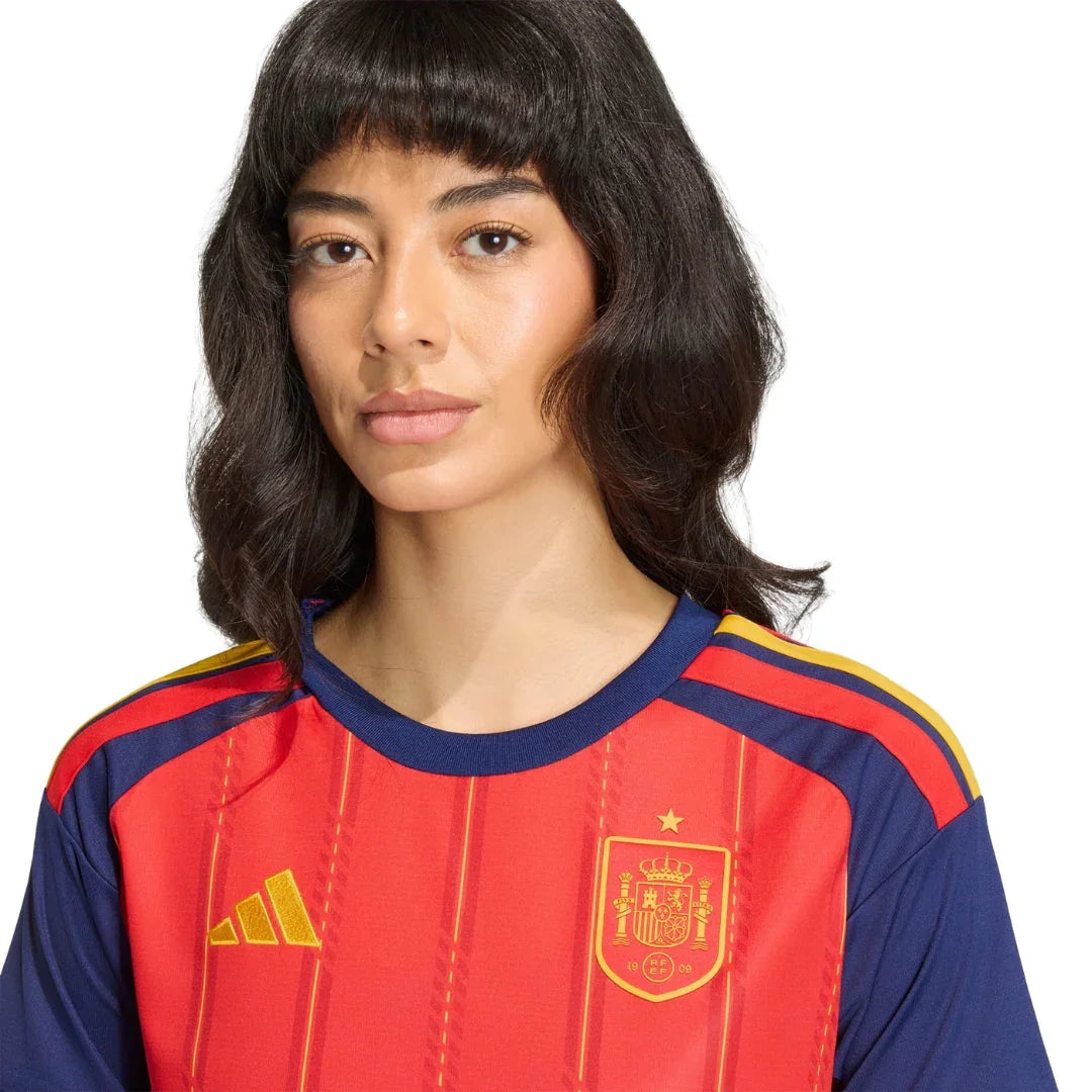 Mujer con camiseta roja de la selección española de fútbol femenino, detalles amarillos y escudo.