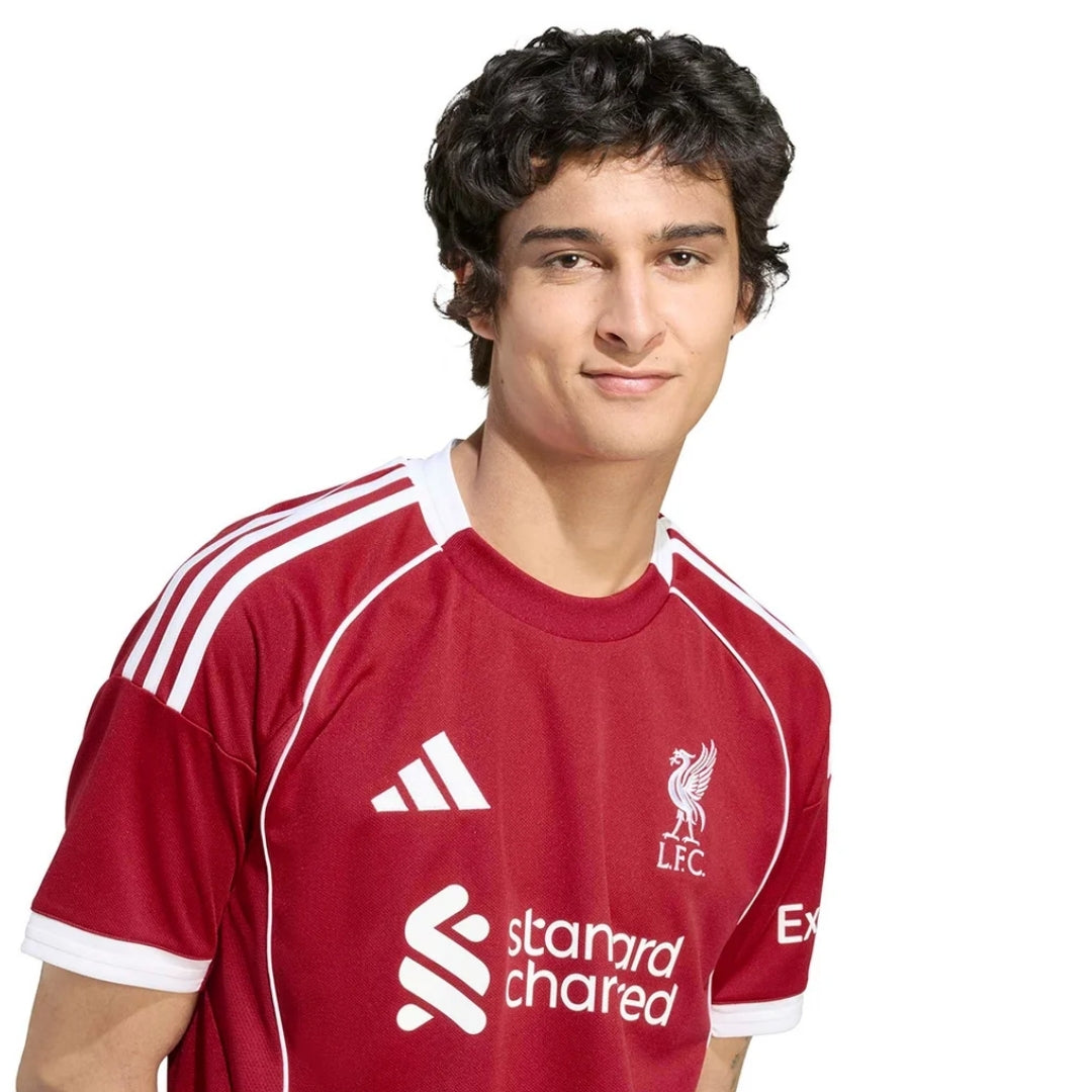 Hombre con camiseta Liverpool 2025 2026 local roja, logos Adidas y Standard Chartered visibles