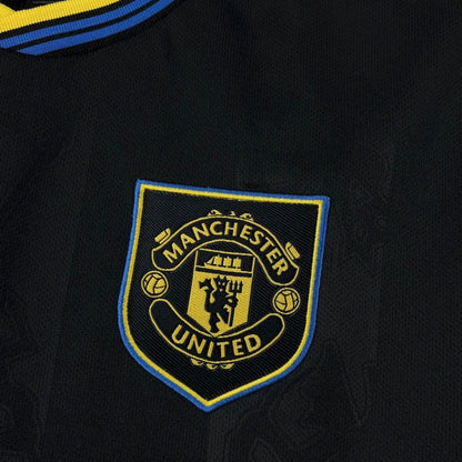 Escudo Manchester United 2025/26 en camiseta negra tercera equipación, detalles azul y amarillo