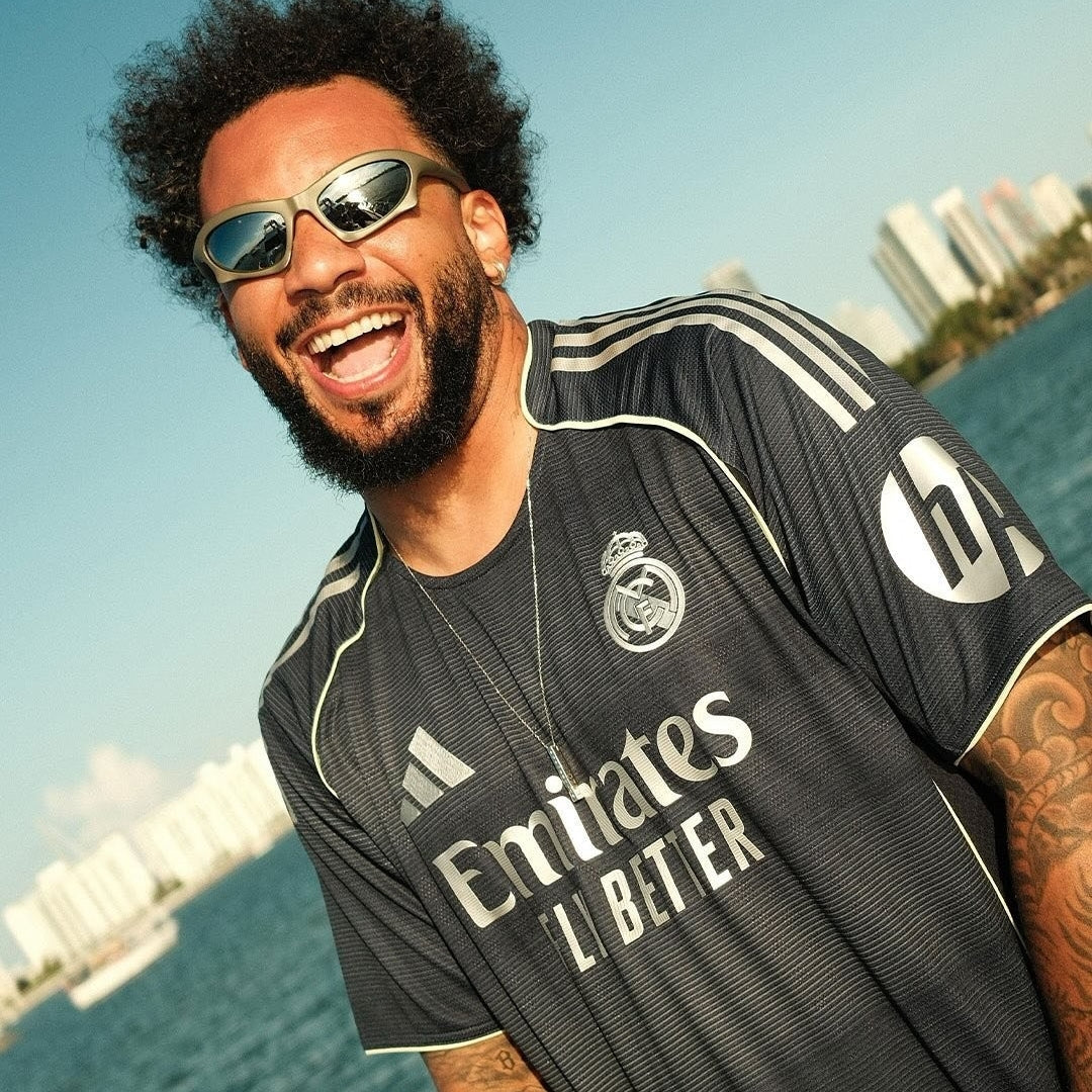 Hombre sonriente con la camiseta negra de visitante del Real Madrid 2025/2026 junto al mar, edificios al fondo