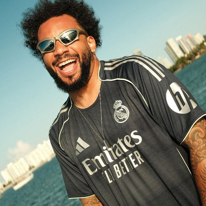 Hombre sonriente con la camiseta negra de visitante del Real Madrid 2025/2026 junto al mar, edificios al fondo