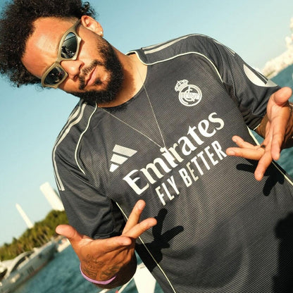Hombre con gafas de sol luciendo la camiseta negra Real Madrid visitante 2025/26, fondo marítimo